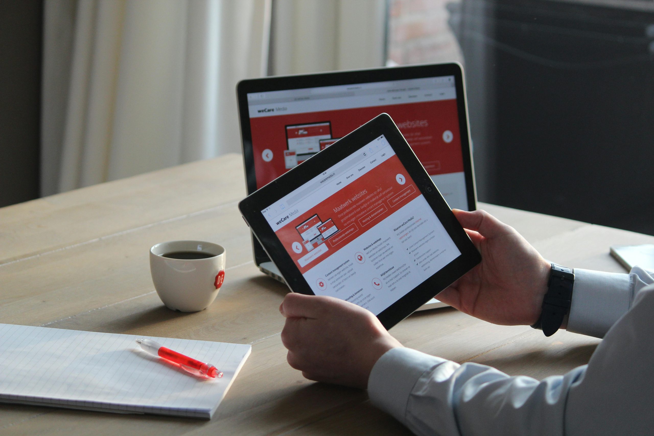 Business professional using a tablet and laptop with a hot drink, focusing on digital content review.
